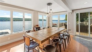 Un vaste espace dédié à la salle à manger, avec une grande table et des chaises en bois, de grandes fenêtres offrant une vue sur un lac et des collines, et un plafond doté d’un éclairage encastré.