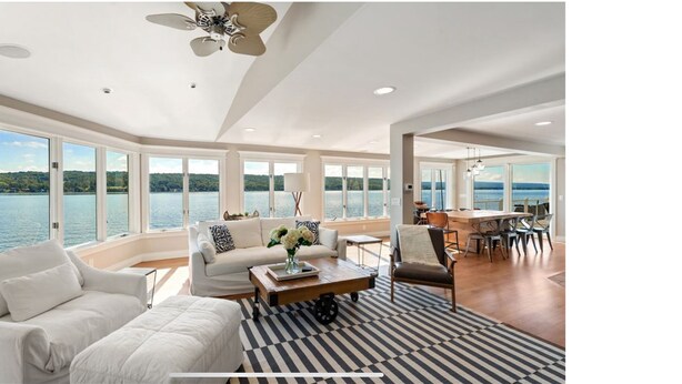 Un vaste espace de vie avec une grande fenêtre donnant sur un lac, un ventilateur de plafond et un coin repas comprenant des chaises et une table.