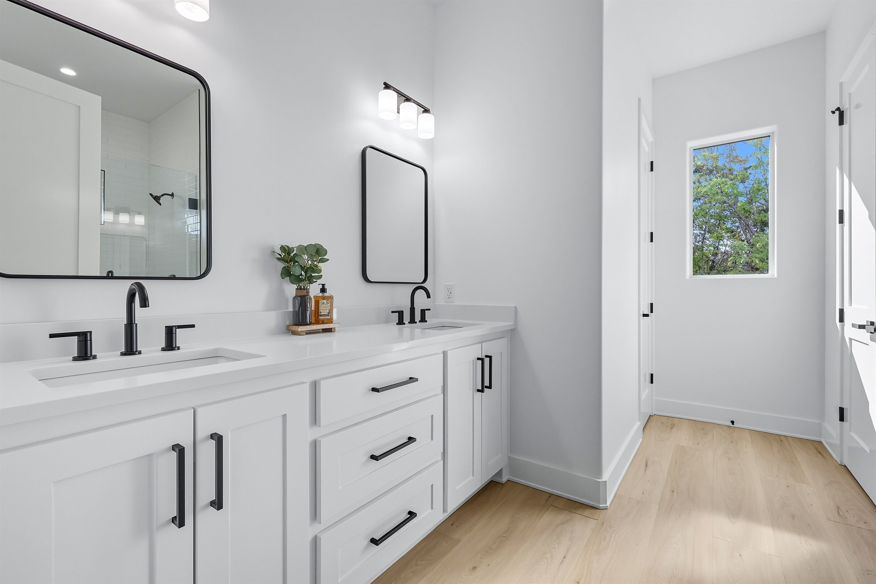 A modern bathroom with a white vanity, two sinks, a large mirror, and a window providing natural light.