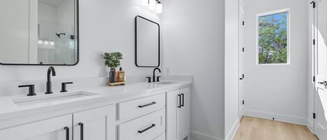 A modern bathroom with a white vanity, two sinks, a large mirror, and a window providing natural light.