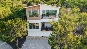 Et moderne hus med to etager, store vinduer og en balkon, omgivet af frodig vegetation.