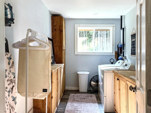 A compact laundry room with wooden cabinets, a washing machine, a dryer, and a window with a view of trees. - The Fern, Huge Spa-Like Home Near Lake Crescent/Sol Duc • Olympic National Park (Port Angeles)