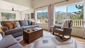 Ein großzügiges Wohnzimmer mit grauem Sofa, einem Holztisch und einem Schaukelstuhl. Große Fenster bieten Blick auf eine Berglandschaft.