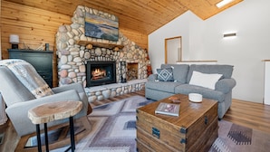 Ein gemütliches Wohnzimmer mit Kamin aus Stein, einem grauen Sofa, einem Holztisch und einem rustikalen Holzstuhl.