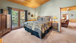 A bedroom with a large bed, wooden ceiling, a wooden cabinet, a desk with a lamp, and a view through a sliding door to an outdoor deck. - The Getaway Chalet - Walk to Slopes & Golf Course, Ping Pong, Fireplace, Deck (Big Bear Lake)