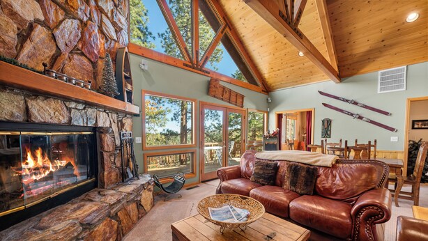 A cozy living room with a stone fireplace, leather sofa, and a dining area with wooden furniture. - The Getaway Chalet - Walk to Slopes & Golf Course, Ping Pong, Fireplace, Deck (Big Bear Lake)