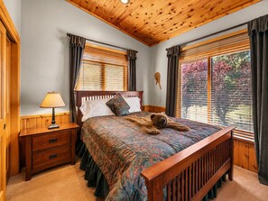 A bedroom with a wooden headboard, a bed with a patterned bedspread, a bedside lamp, and a window with blinds.