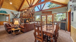 A cozy lodge interior with a stone fireplace, wooden furniture, and large windows offering views of the outdoors. - The Getaway Chalet - Walk to Slopes & Golf Course, Ping Pong, Fireplace, Deck (Big Bear Lake)