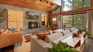 A spacious living room with a stone fireplace, wooden ceiling, and large windows offering a view of trees. - Modern Primrose - Spacious 3 BR Mountain Cabin | Decks, A/C, Game Room & Near Ski, Zoo & Golf (Big Bear Lake)