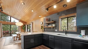 A modern kitchen with dark cabinets, a wooden ceiling, and a large window. - Modern Primrose - Spacious 3 BR Mountain Cabin | Decks, A/C, Game Room & Near Ski, Zoo & Golf (Big Bear Lake)