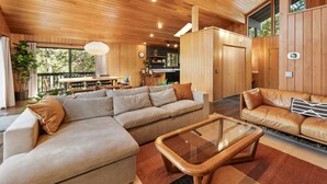 A spacious living room with a large sectional sofa, a smaller sofa, and a glass coffee table. There is a dining area with a table and chairs, and a kitchen area with wooden cabinets. - Modern Primrose - Spacious 3 BR Mountain Cabin | Decks, A/C, Game Room & Near Ski, Zoo & Golf (Big Bear Lake)
