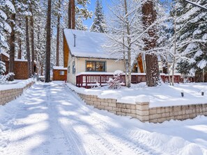 A cabin with a snow-covered roof, surrounded by trees, and a brick pathway leading to the entrance. - Peregrine Chalet - Hot Tub + Fenced Yard | Walk to Bear Mtn (Big Bear Lake)