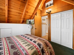 A bedroom with a wooden ceiling, a bed with a patterned bedspread, a television on the wall, and a white door.