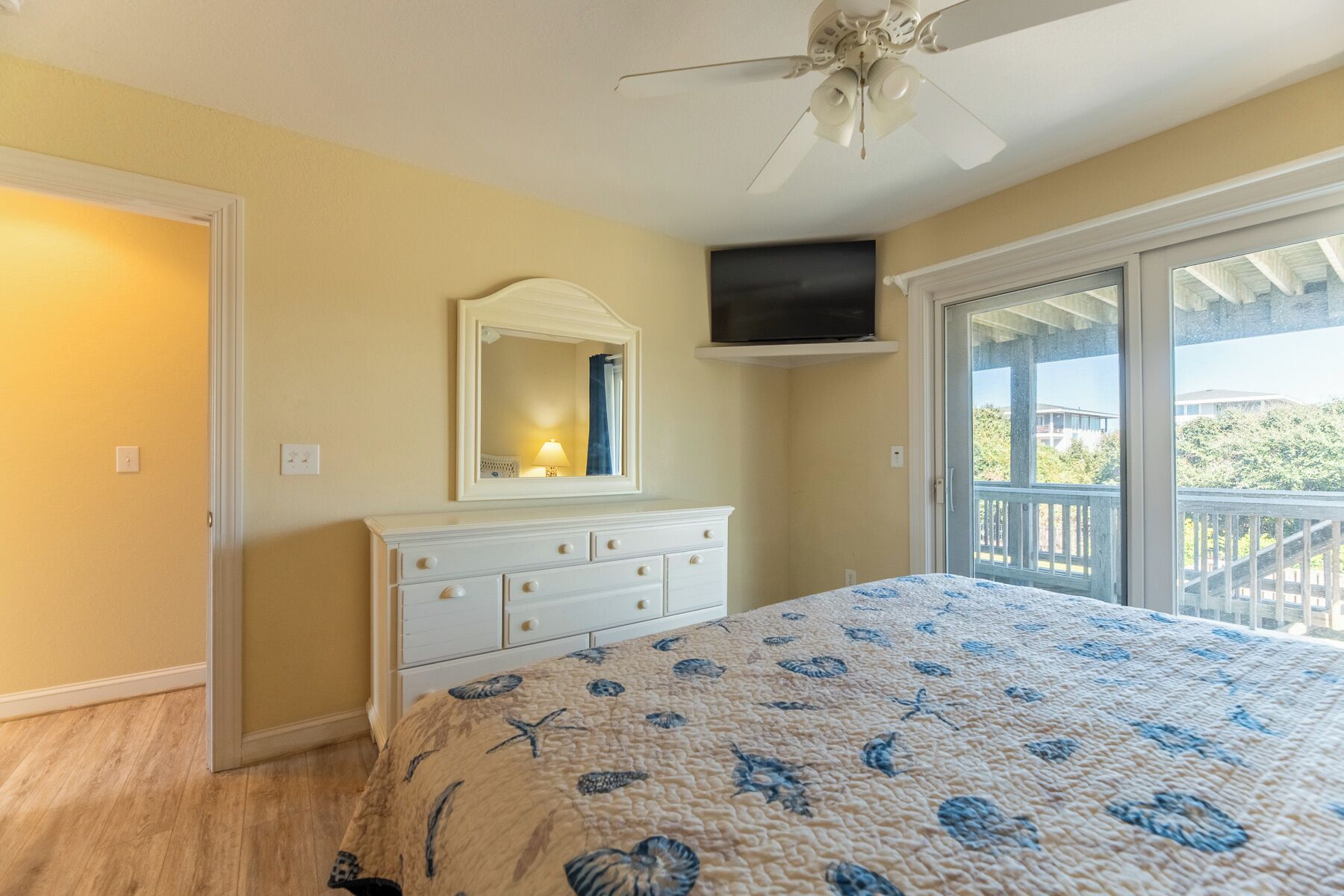 Quarto com cama, cômoda, ventilador de teto, televisão e varanda com vista.