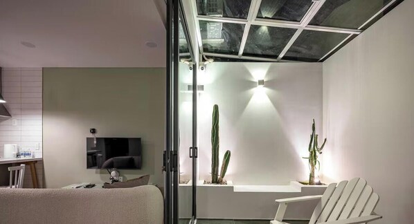 Un soggiorno moderno con parete in vetro, televisore a schermo piatto, una poltrona bianca e una pianta di cactus.