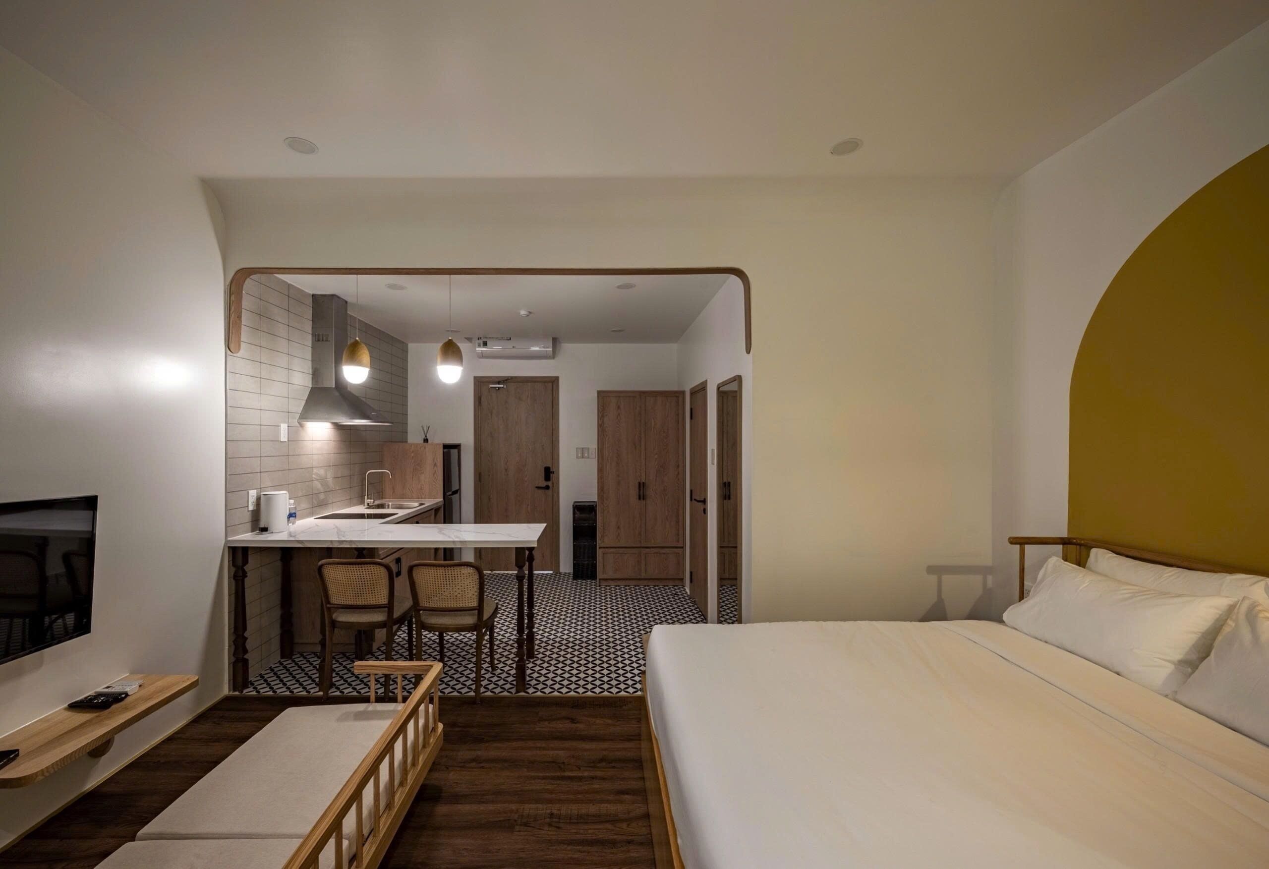 A compact hotel room with a bed, a small desk with a chair, a TV, and a kitchenette.