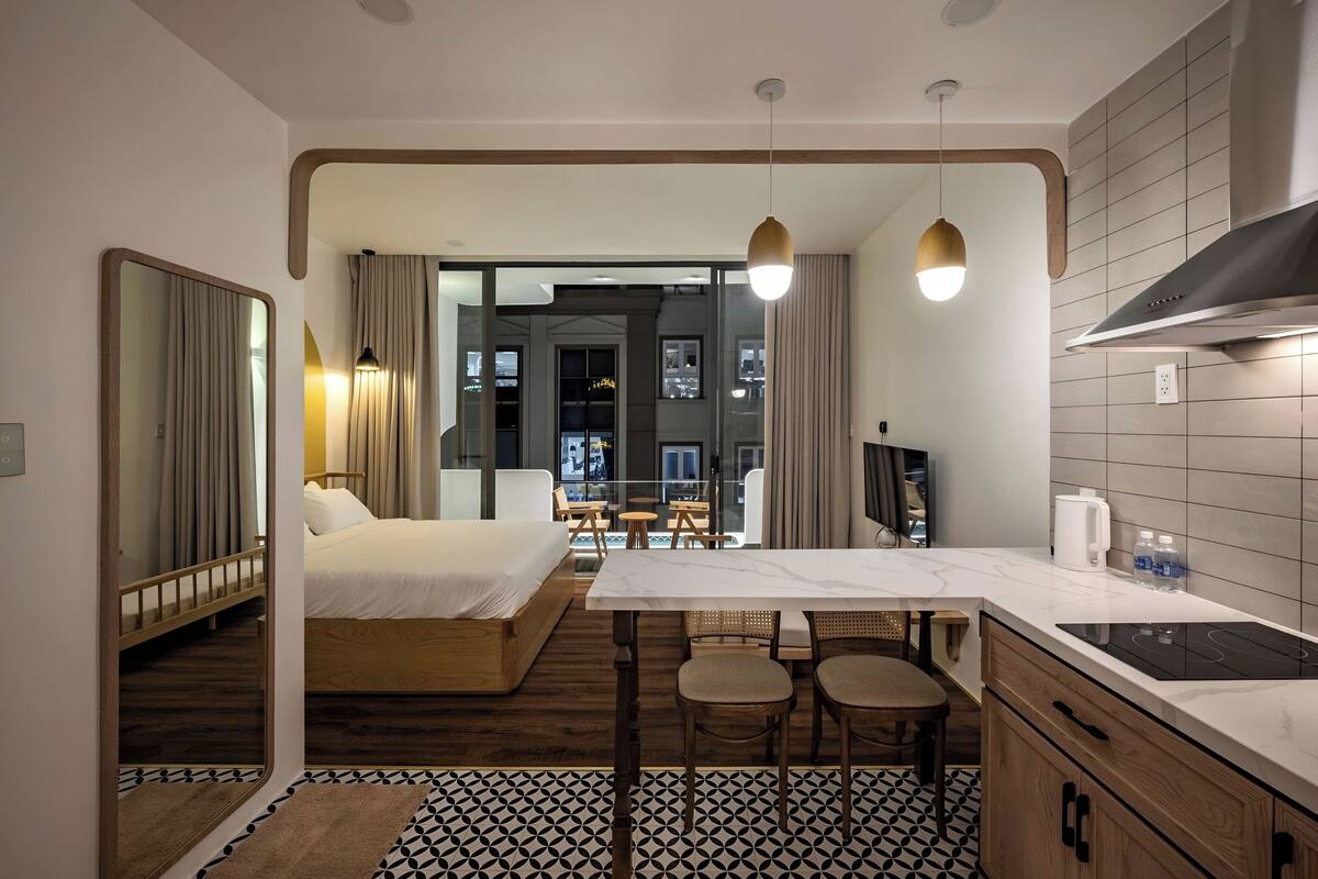 A modern hotel room with a bed, a dining area with stools, a kitchen with a stove, and a large mirror.