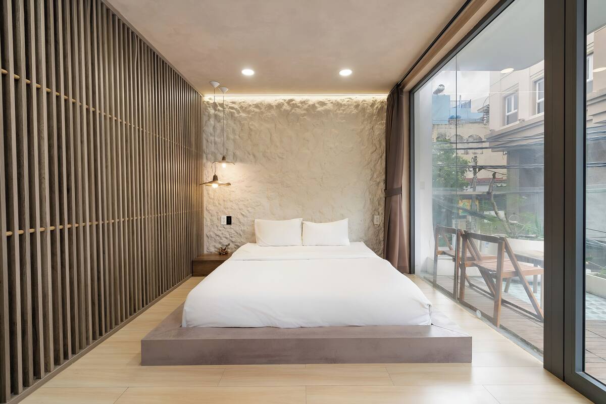 A modern bedroom with a large bed, a wooden slat wall, a stone-textured wall, and a view of a balcony with outdoor furniture.