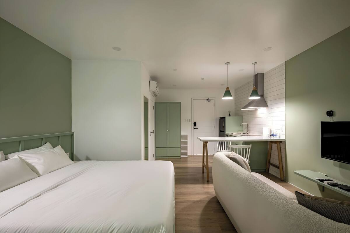 A modern hotel room with a large bed, a sofa, a dining area, and a kitchen.