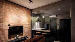 Una moderna camera d'albergo con parete decorata in mattoni, televisione a schermo piatto, divano in pelle, angolo cottura con lavandino e sgabelli da bar, e un grande letto.