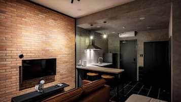 Une chambre d’hôtel moderne avec un mur d’accent en briques, un téléviseur à écran plat, un canapé en cuir, un petit coin cuisine avec un évier et des tabourets de bar, et un grand lit.