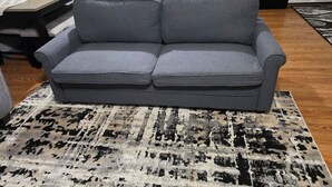 Ein graues Sofa mit schwarzem Akzent, ein gemustertes Teppich, ein Esstisch mit Stühlen und eine Küchenzeile im Hintergrund.