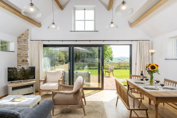 A spacious living room with a television, comfortable seating, a dining area with a table set for four, and a large sliding glass door leading to a garden.