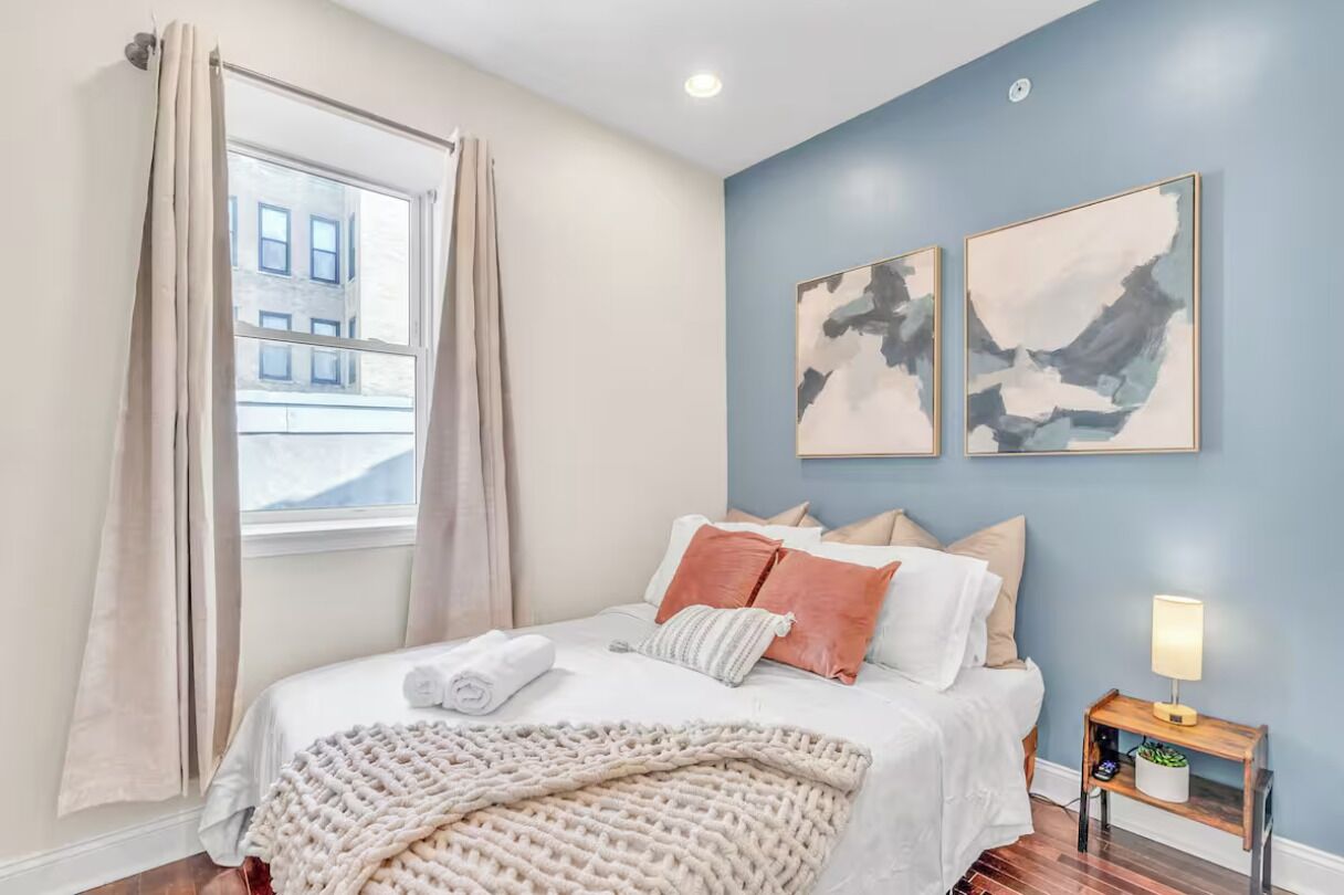 A bedroom with a large bed, beige curtains, a window with a view of buildings, and two abstract wall art pieces.