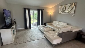 A living room with a sectional sofa, a flat-screen TV, a sliding glass door, and a wall-mounted artwork. - 3-bedroom house in welcoming Port Charlotte with AC (Port Charlotte)