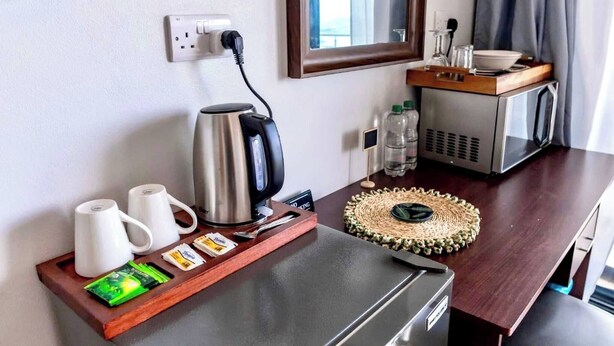 Habitación de hotel con kitchenette, cafetera, tetera, microondas, refrigerador pequeño, bandeja con tazas de café y bolsitas de té, posavasos tejido y espejo sobre la encimera.