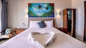 Una habitación de hotel con una cama, una toalla con forma de corazón, un cuadro de una escena tropical, una mesita de noche, una silla y un armario.