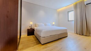 A modern bedroom with a large bed, wooden flooring, and a nightstand with a lamp. - Han Dhahrat Laban (Riyadh)