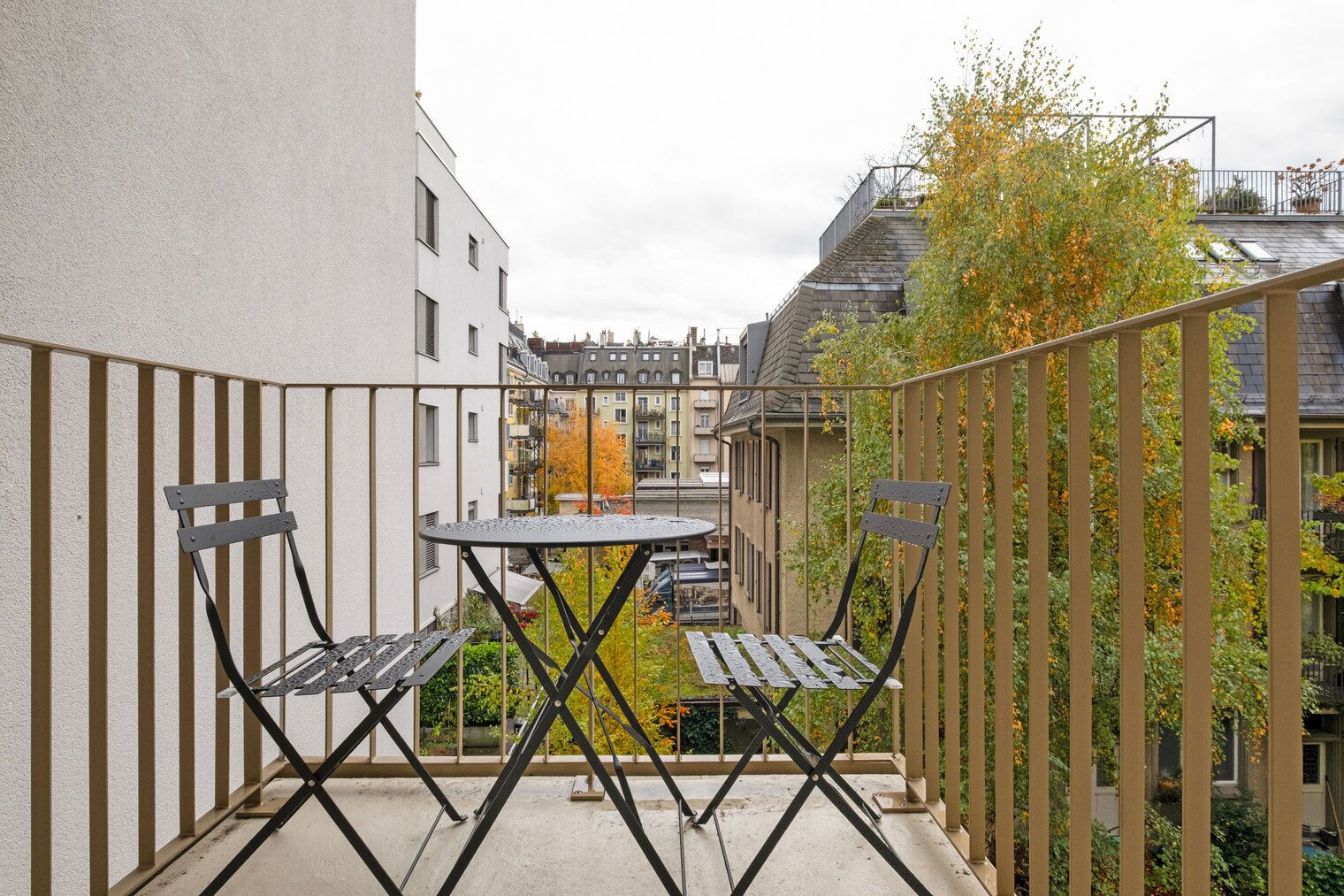 Ein Balkon mit Tisch und zwei Stühlen, umgeben von einem Metallgeländer, mit Blick auf ein Wohngebiet im Herbstlaub.
