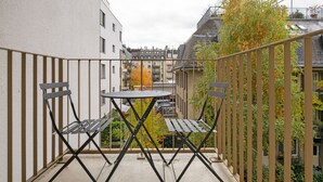 Ein Balkon mit Tisch und zwei Stühlen, umgeben von einem Metallgeländer, mit Blick auf ein Wohngebiet im Herbstlaub.