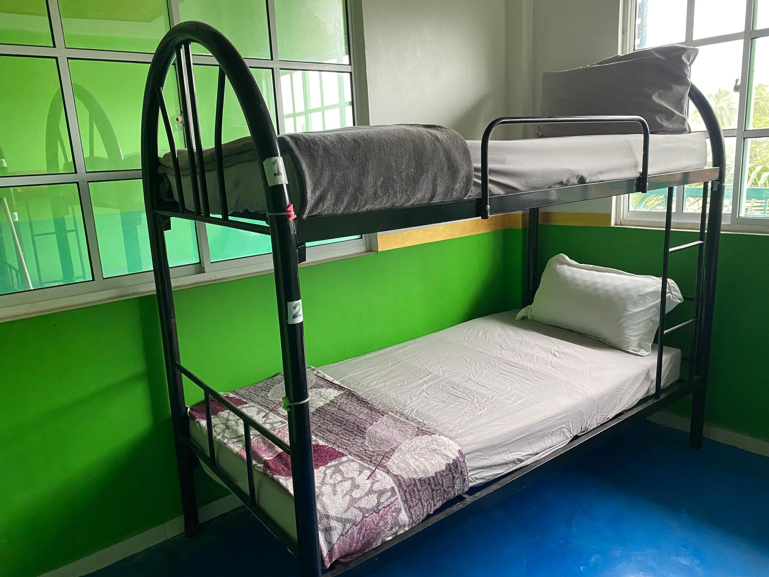 Ein Etagenbett mit grauer Decke und weißem Kopfkissen, eine gemusterte Decke auf dem unteren Bett sowie ein Fenster mit grünen Wänden und Blick ins Grüne.