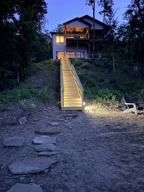 A wooden staircase leading to a modern house with a balcony, surrounded by trees.