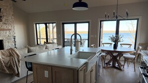 A modern kitchen with a central island, large windows with a view of the sea, and a dining area with wooden furniture.