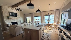 A modern kitchen with a central island, a dining area with a wooden table and chairs, a built-in oven, and a stone fireplace.