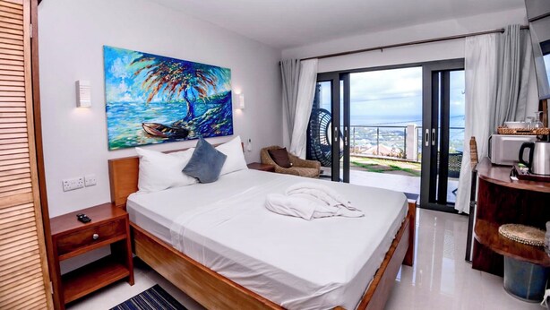Ein Hotelzimmer mit Bett, einem Gemälde einer tropischen Szene, Meerblick und einem Balkon mit Stuhl.