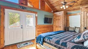 Un dormitorio tipo cabaña con cama, ventilador de techo, televisor y puerta con ventana.