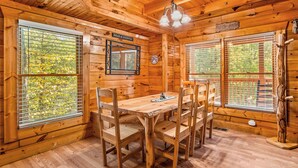 Interior de una cabaña de madera con una mesa de comedor y sillas, ventanas grandes con persianas y una lámpara colgante.