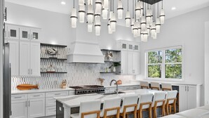 A modern kitchen with white cabinetry, a central island with bar stools, a large window with a view of trees, and a unique light fixture. - Villa Rosa - Coastal Home Complete with Private Pool and Outdoor Dining (Santa Rosa Beach)