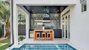 A modern outdoor kitchen with a pool, featuring a stone backsplash, wooden furniture, and a ceiling fan. - Villa Rosa - Coastal Home Complete with Private Pool and Outdoor Dining (Santa Rosa Beach)