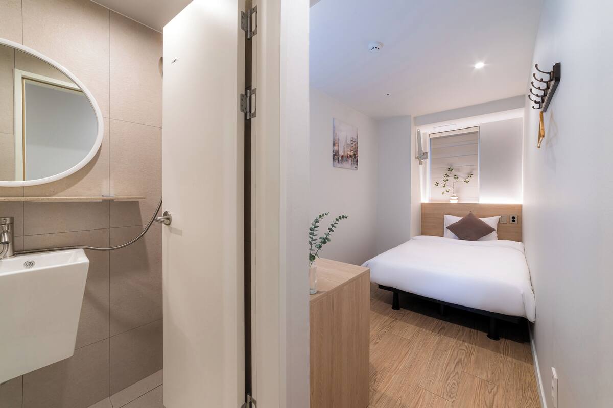 A modern hotel room with a wooden floor, a bed with white bedding, a bedside table with a vase, a wall-mounted mirror, and a small picture.