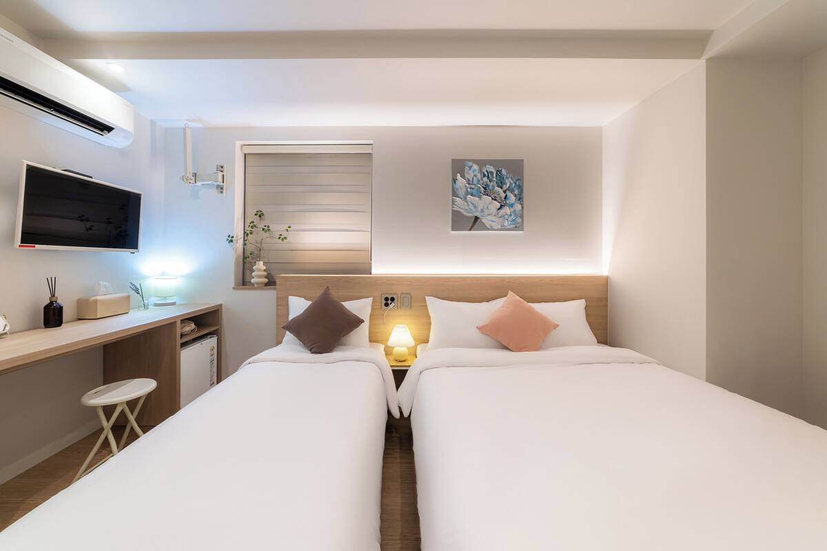 A modern hotel room with two single beds, a desk with a chair, a TV, and a small table with a lamp.