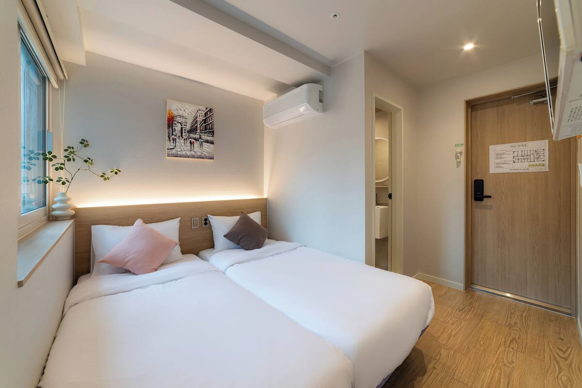 A double bed with white bedding and two pillows, a wall-mounted air conditioner, a framed picture, and a wooden door.