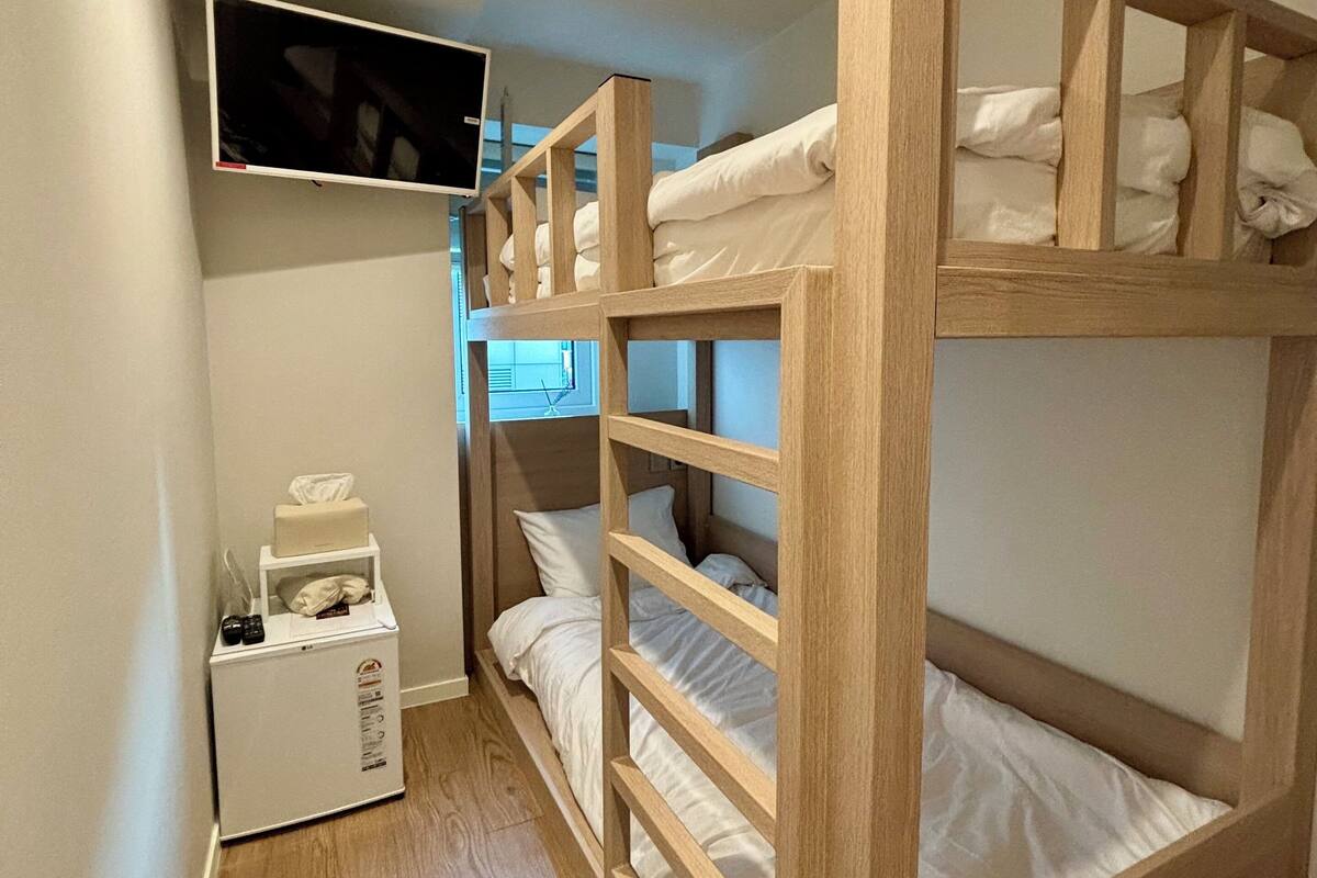 A bunk bed room with a television mounted on the wall, a small white cabinet, and wooden flooring.