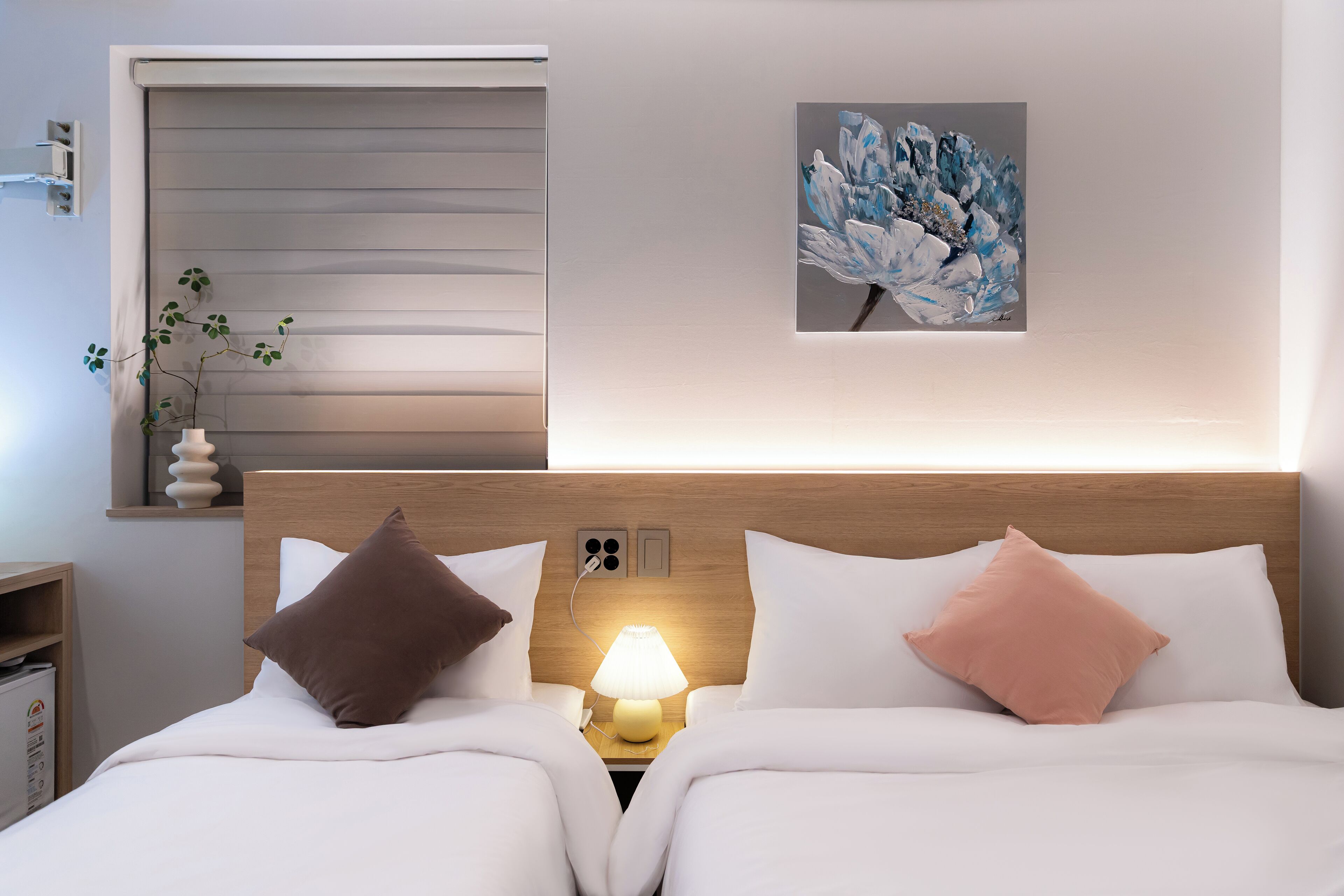 A hotel room with two beds, a bedside table with a lamp, a wall-mounted artwork, and a window with blinds.