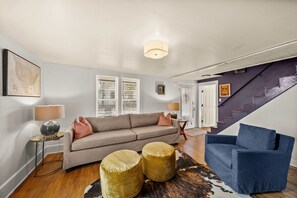 Un salon avec un canapé, des poufs, un fauteuil bleu, une lampe et une carte accrochée au mur.