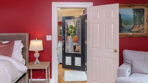 A hotel room with a bed, a bedside table with a lamp, a door leading to a hallway, and a painting on the wall. - Eureka Springs 3 Bed - Teacup Cottage (Eureka Springs)